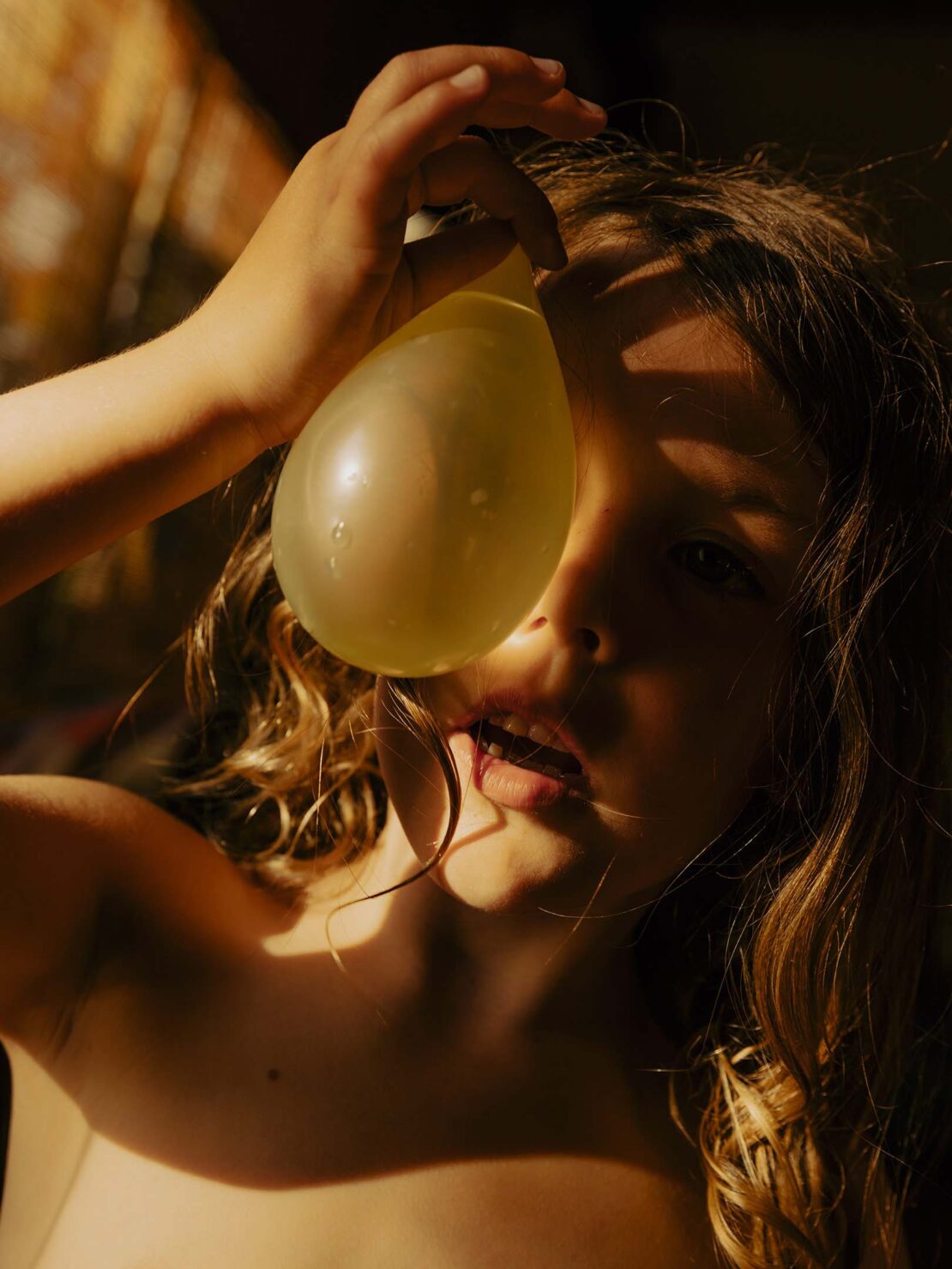 Close-up portrait of a young child holding a water balloon to their face in a warm, sunlit indoor setting. The child has curly hair.
