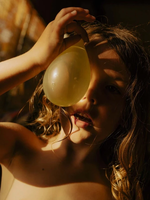 Close-up portrait of a young child holding a water balloon to their face in a warm, sunlit indoor setting. The child has curly hair.