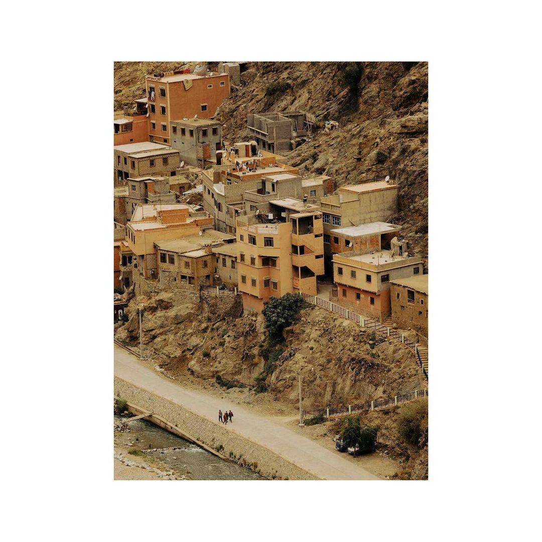 Hillside village in Morocco with earth-tone buildings clustered together on a rocky slope, a road, and three people walking along the path.