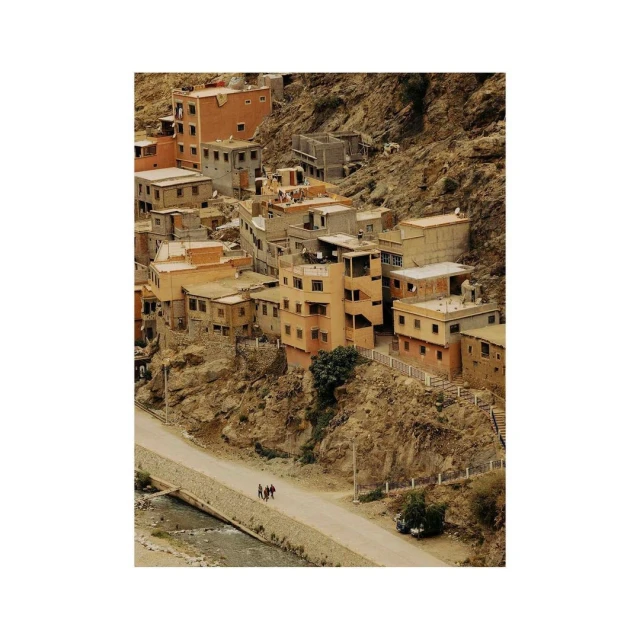 Hillside village in Morocco with earth-tone buildings clustered together on a rocky slope, a road, and three people walking along the path.