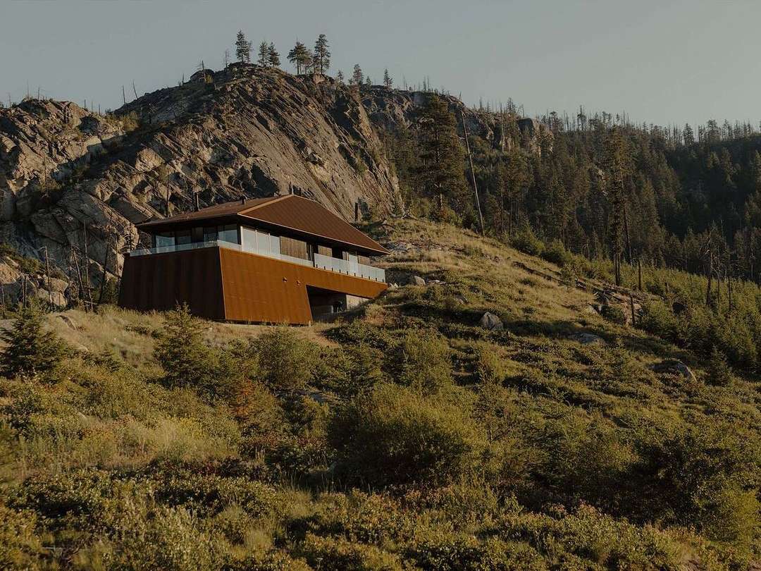 Exterior of a modern, angular house set on a grassy hillside with a rocky mountain in the background, showcasing architectural design.