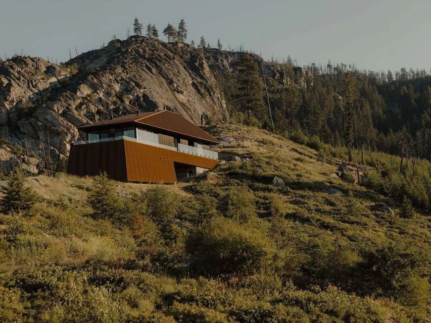 Exterior of a modern, angular house set on a grassy hillside with a rocky mountain in the background, showcasing architectural design.