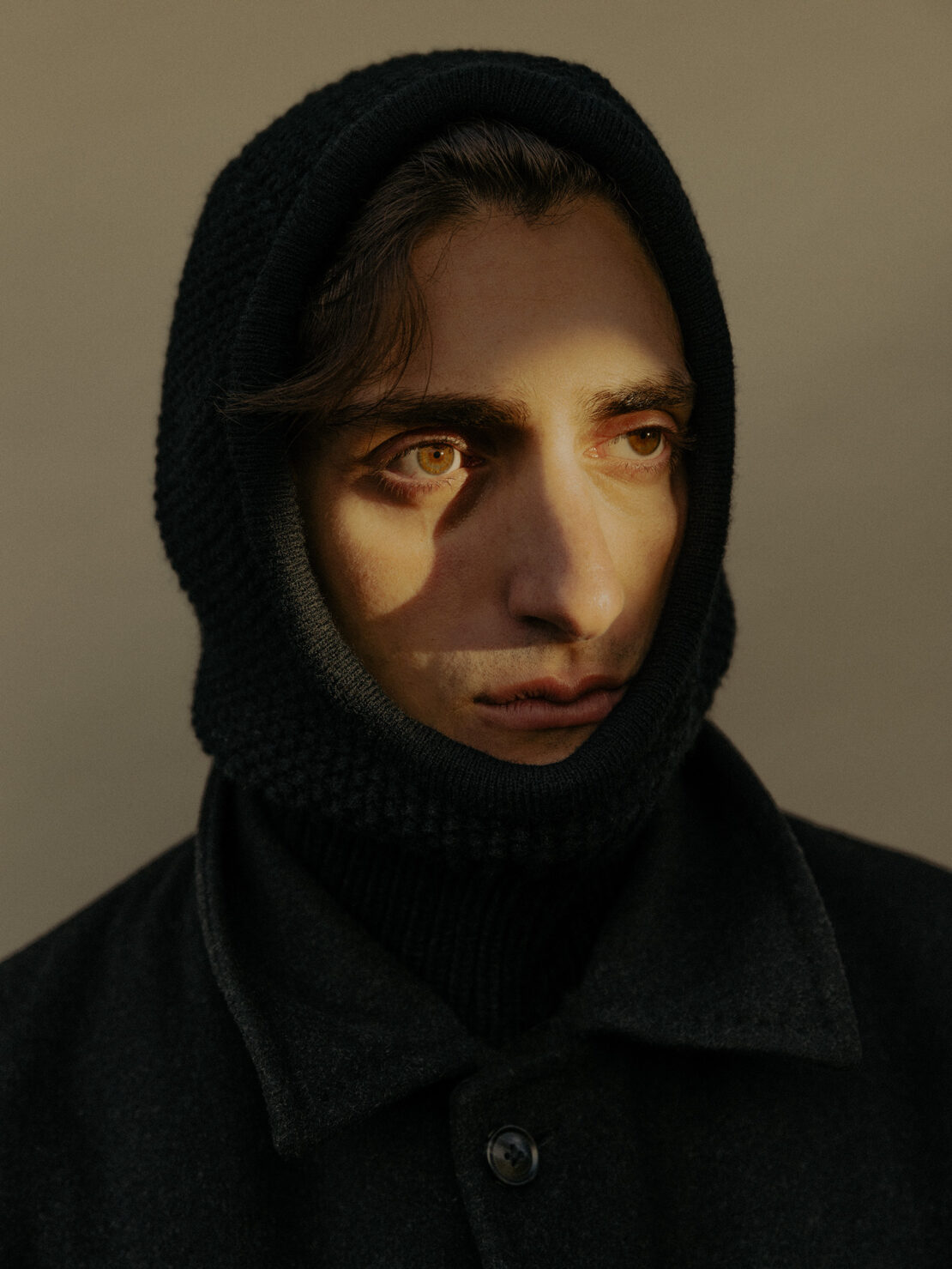 Close-up portrait of a young adult male with dark hair and olive skin wearing a black knit hood and black jacket in studio lighting.