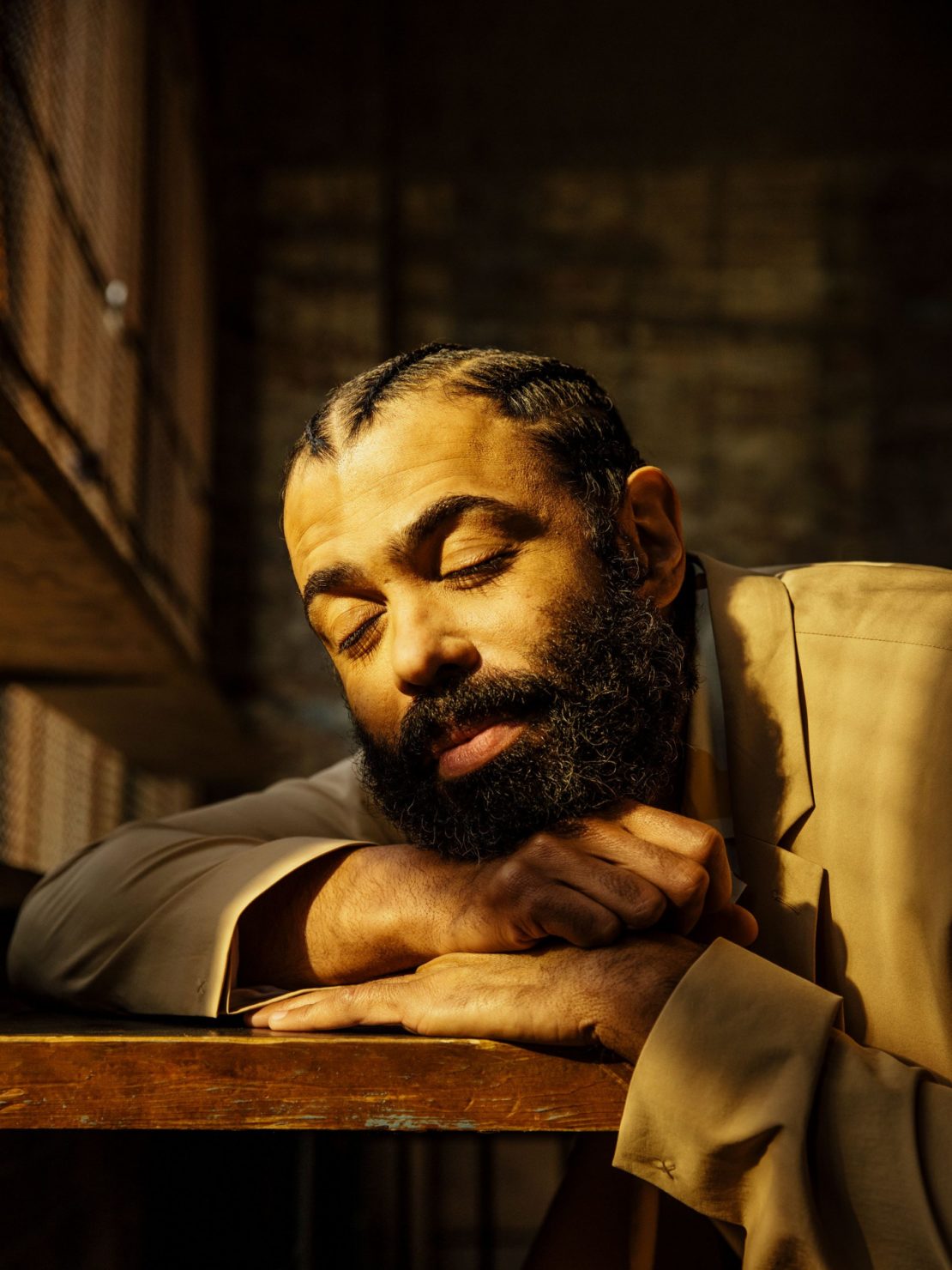 Portrait of an adult man with a beard and cornrow braids, wearing a tan suit, with eyes closed and arms resting on a dark wooden surface.