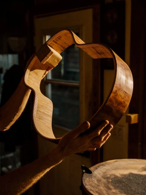 Unfinished guitar body held by a person in a warm, wood-paneled workshop with natural light illuminating the instrument's structure.