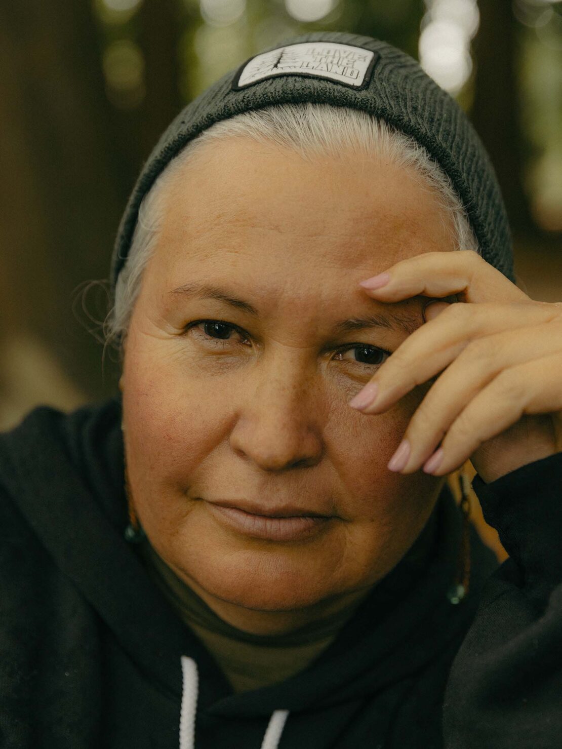 Close-up portrait of a mature adult female with grey hair, wearing a grey beanie and a dark hoodie outdoors with a blurred forest background.