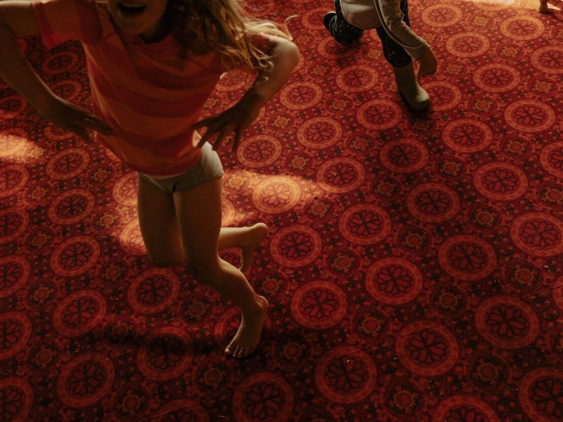 Young girl jumping indoors on red patterned carpet, expressing joy and movement, captured from a high angle.