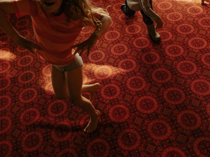 Young girl jumping indoors on red patterned carpet, expressing joy and movement, captured from a high angle.