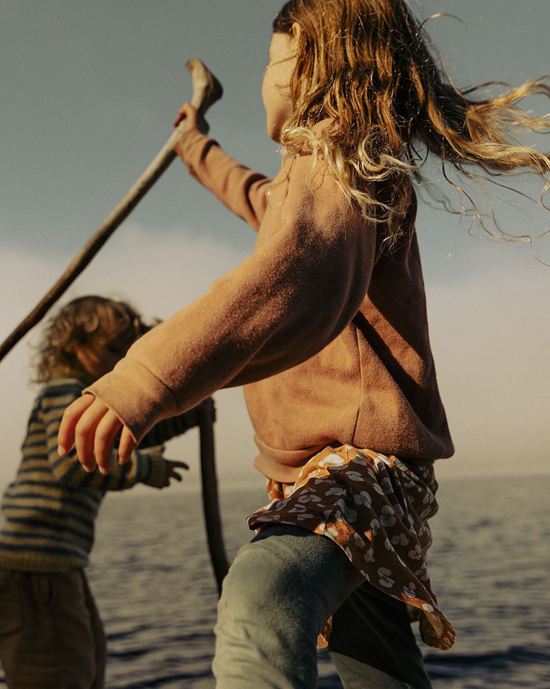 Two children are playing on the water's edge with a stick. One wears a striped shirt, and the other has on a rust-colored sweatshirt and jeans.