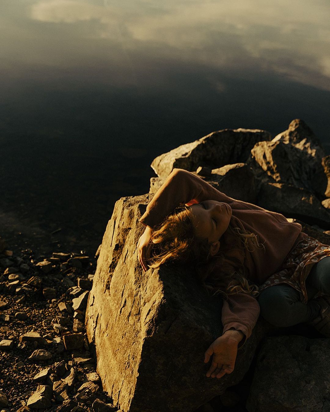 Young woman with long hair rests on a rock in an outdoor setting, showcasing a serene pose against a backdrop of distant mountains.