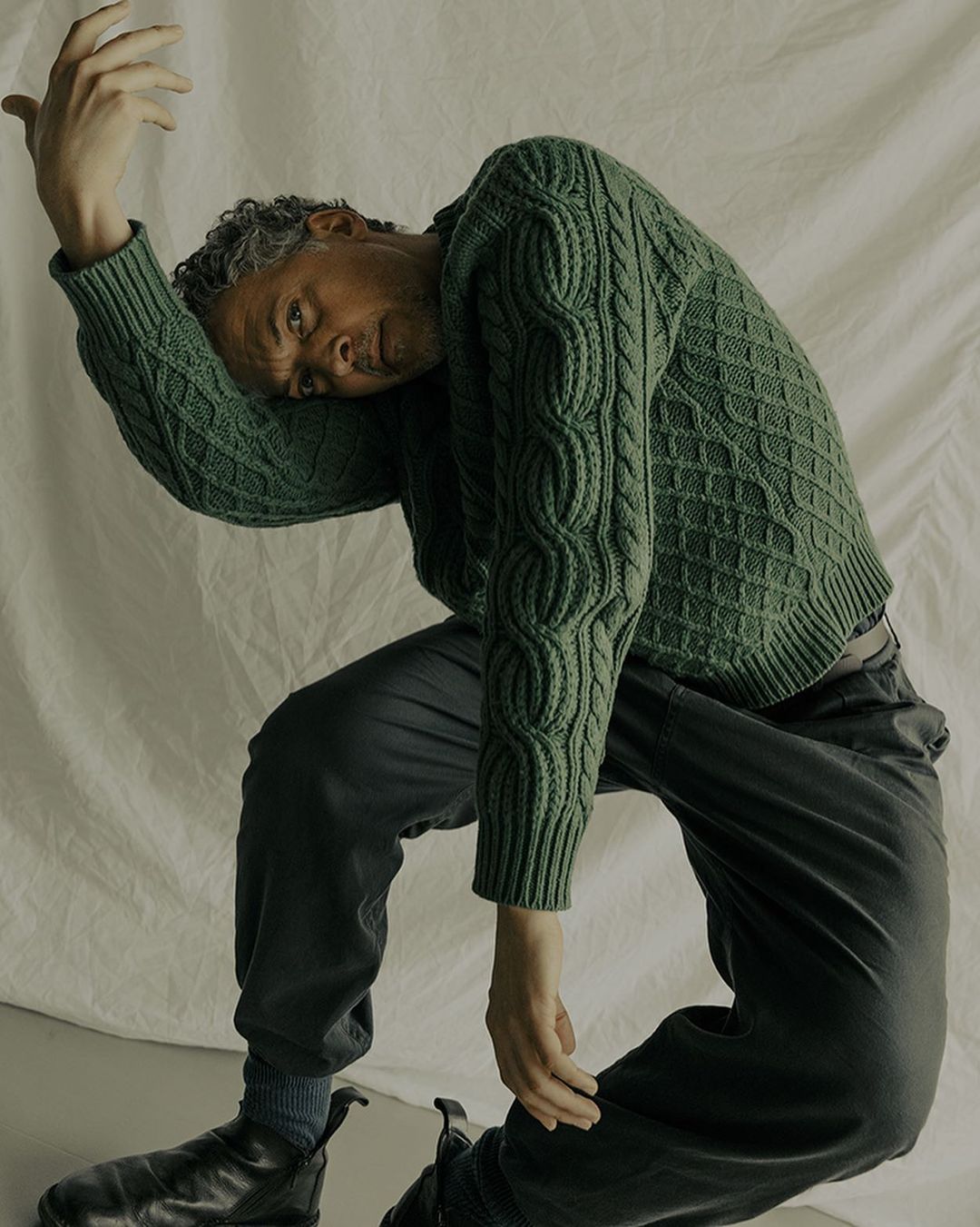Man in green cable-knit sweater poses kneeling on floor in photo studio with white backdrop, fashion style and portrait-style image.