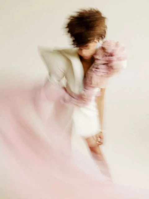 Motion blur fashion shot of a woman with dark hair wearing a white dress with a large, pink, ruffled shoulder piece in a studio.