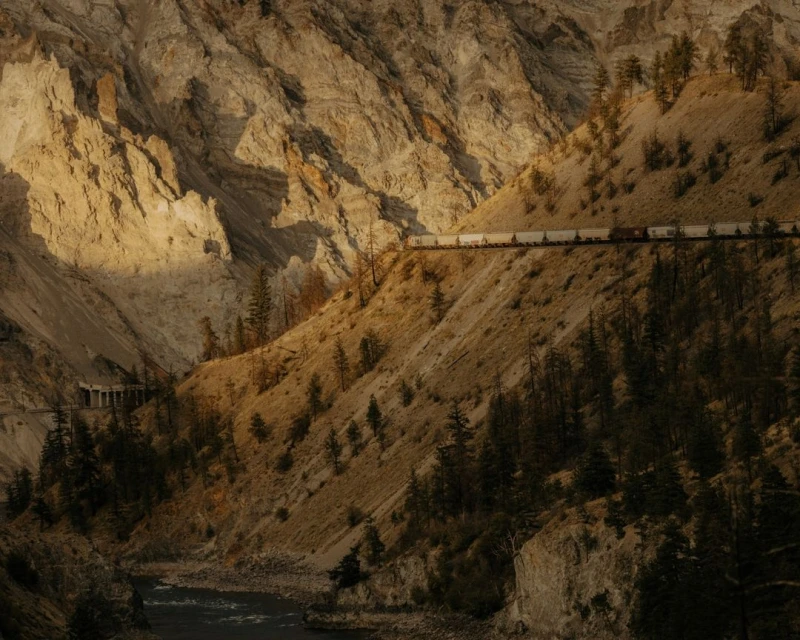 Train winds through a canyon, cutting through the landscape with towering rock formations, trees, and a flowing river in the foreground.