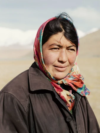 Woman wearing a colorful headscarf and a dark coat in a mountain landscape with grassy plains and snow-capped peaks on the horizon.
