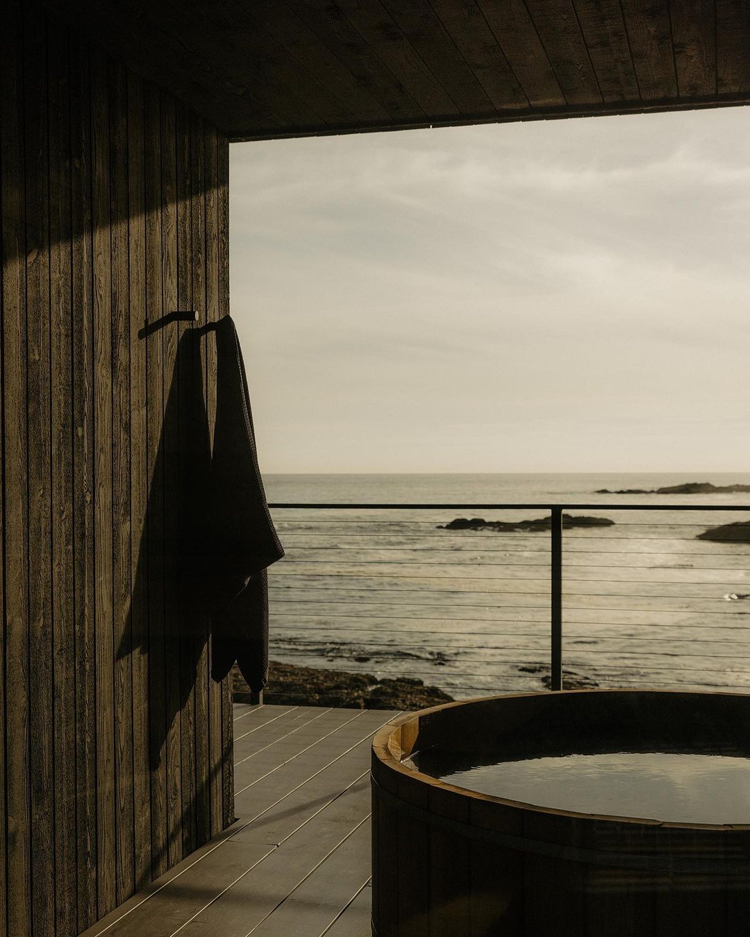Minimalist coastal cabin scene with a dark towel hanging near a wood-paneled hot tub overlooking the ocean horizon from a covered deck.