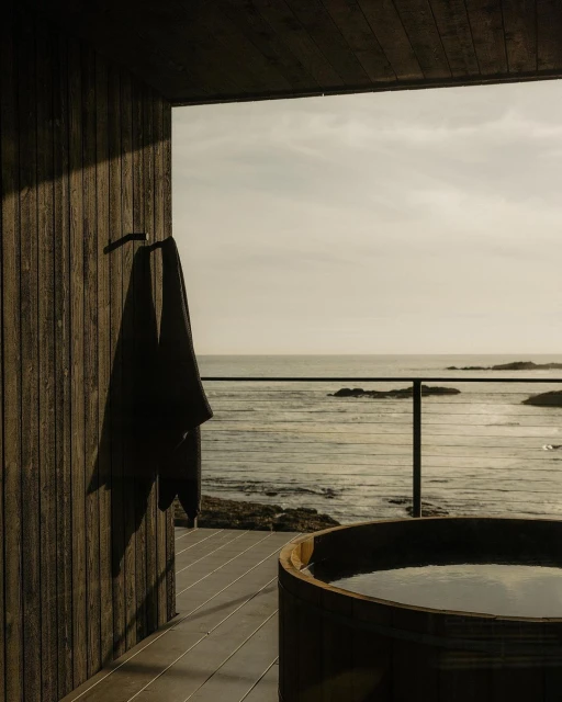 Minimalist coastal cabin scene with a dark towel hanging near a wood-paneled hot tub overlooking the ocean horizon from a covered deck.