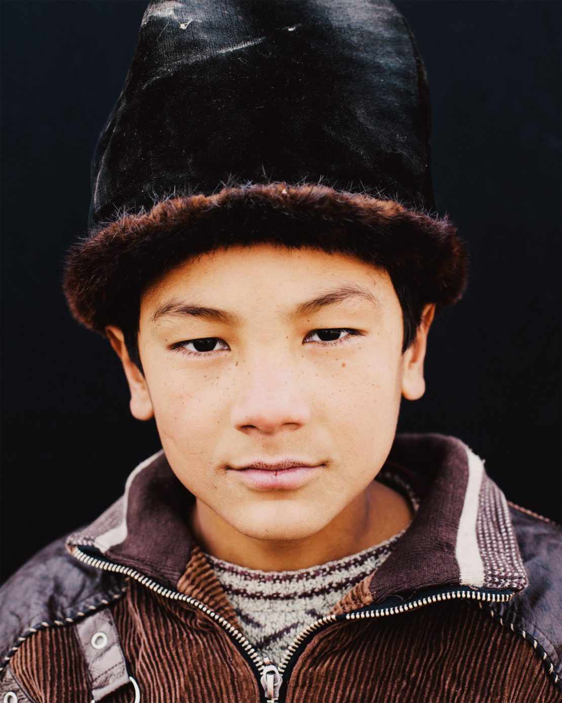 Portrait of a young person wearing a traditional hat with fur trim, looking directly at the camera against a dark background.