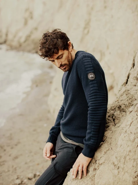 Adult male poses next to a rock formation, wearing a navy ribbed wool sweater and dark grey pants against a neutral background.