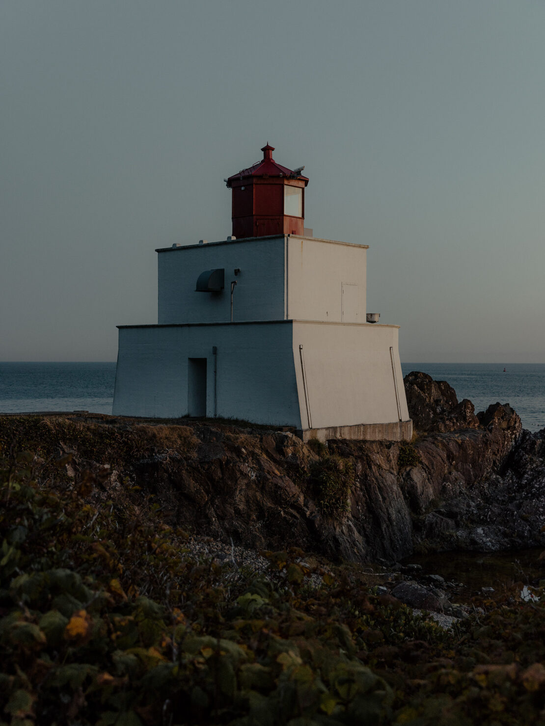 Lighthouse on a rocky cliff at dusk, featuring a two-story white structure with a red lantern room against a tranquil sky and ocean backdrop.