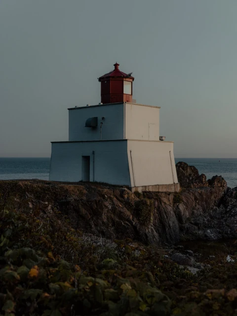 Lighthouse on a rocky cliff at dusk, featuring a two-story white structure with a red lantern room against a tranquil sky and ocean backdrop.