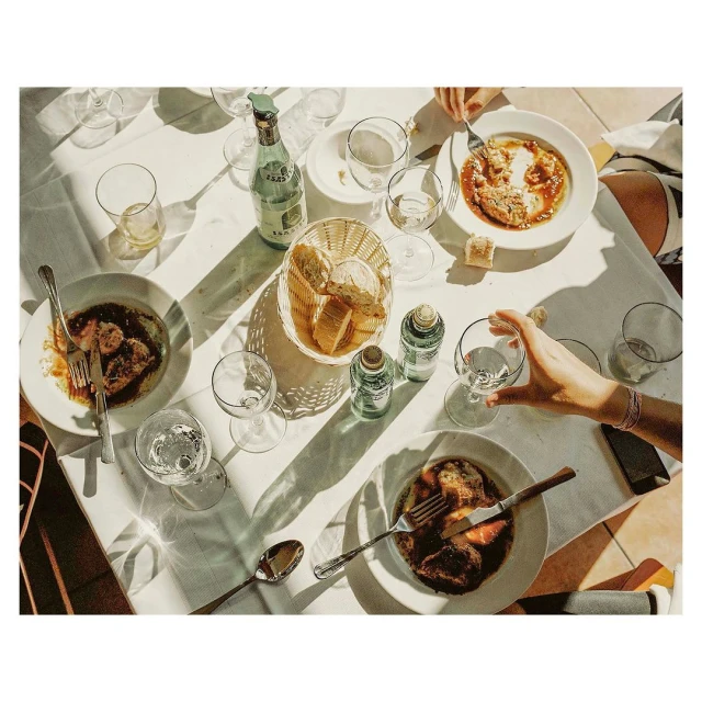 Overhead view of a sunlit outdoor dining table topped with plates of food, bottles of beverages, and glasses of water with a few diners.