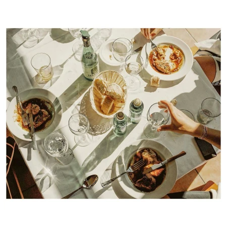 Overhead view of a sunlit outdoor dining table topped with plates of food, bottles of beverages, and glasses of water with a few diners.