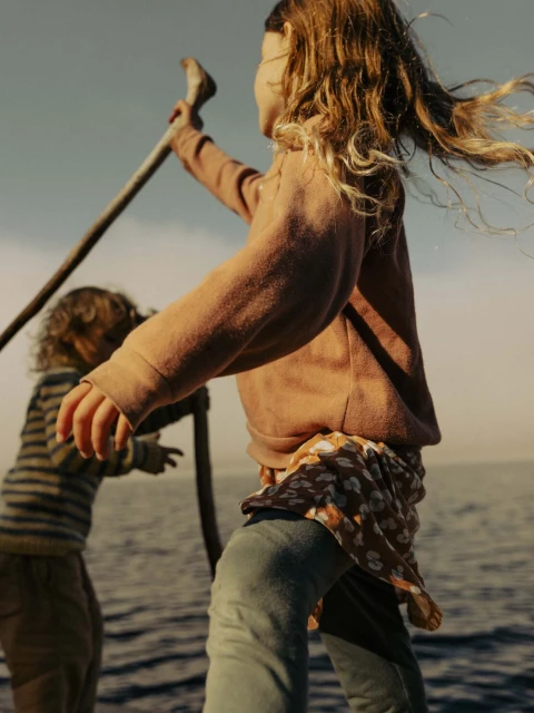 Two children stand on a boat deck with a calm ocean and hazy sky. One child holds a stick in the air, with curly hair blowing in the wind.
