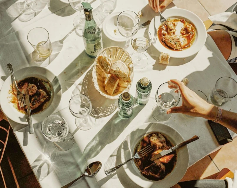 Overhead shot of a white table with plates of food, cutlery, glasses, bottles of water, and bread, some people are visible.