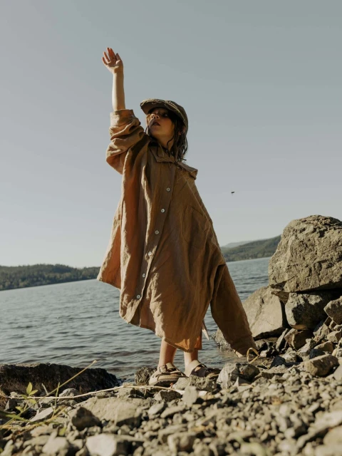 Young child in oversized shirt and cap stands on rocky lake shore, raising arm to sky. Sky is clear and blue, lake water is calm.