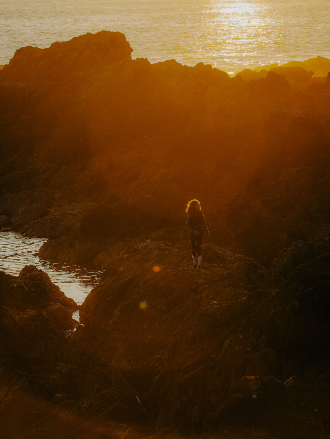 Woman walking on a rocky coast at sunset with golden light reflecting off the ocean water and surrounding rock formations.