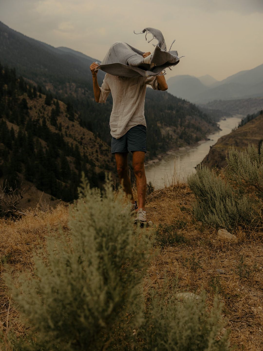 Adult stands with blanket over head, on skateboard, overlooking scenic mountain landscape, captured in medium shot, outdoor setting.