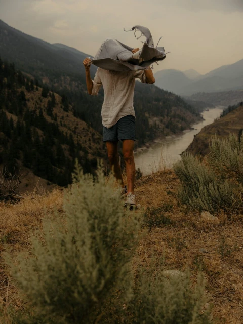 Adult stands with blanket over head, on skateboard, overlooking scenic mountain landscape, captured in medium shot, outdoor setting.