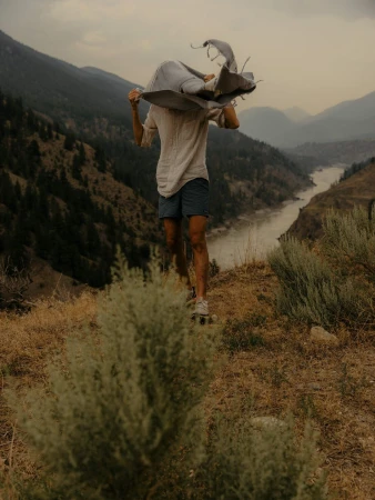 Adult stands with blanket over head, on skateboard, overlooking scenic mountain landscape, captured in medium shot, outdoor setting.