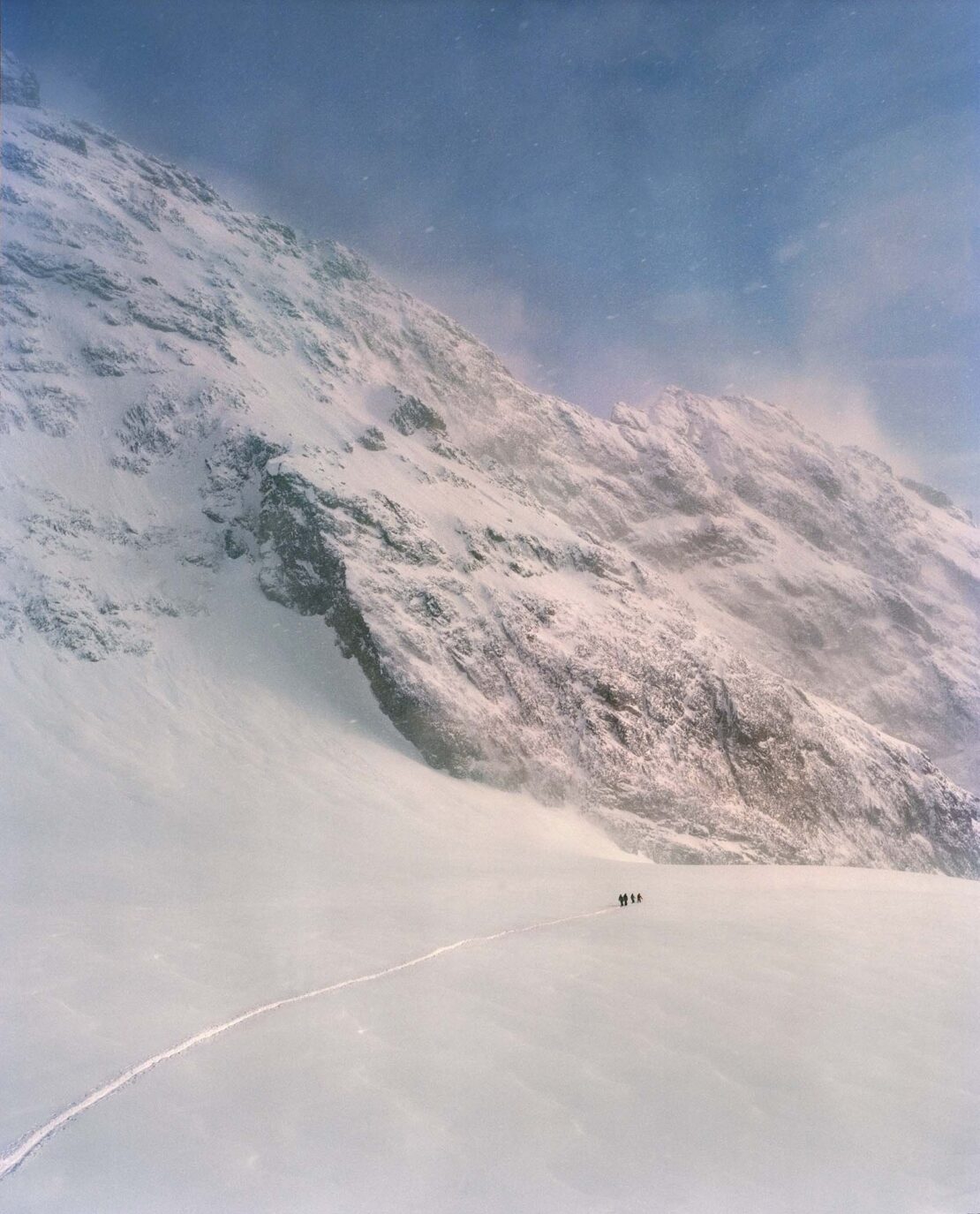 A group of people walking across a snowy plain towards a large, snow-covered mountain under a hazy blue sky with low visibility.