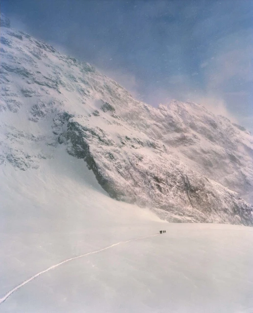 A group of people walking across a snowy plain towards a large, snow-covered mountain under a hazy blue sky with low visibility.