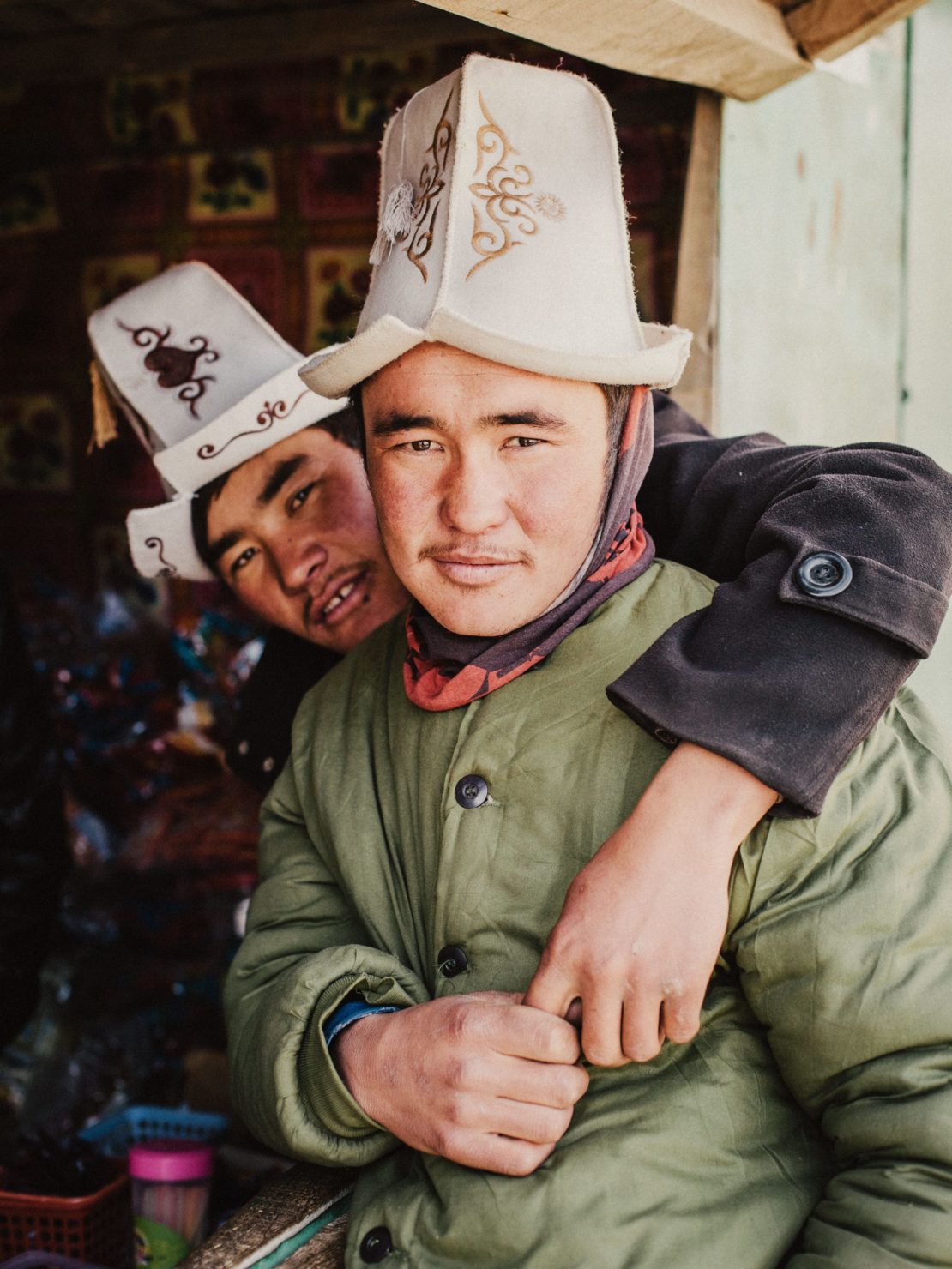 Two adult men wearing traditional Kyrgyz hats and winter jackets pose indoors with warm lighting and neutral color grading in a medium shot.