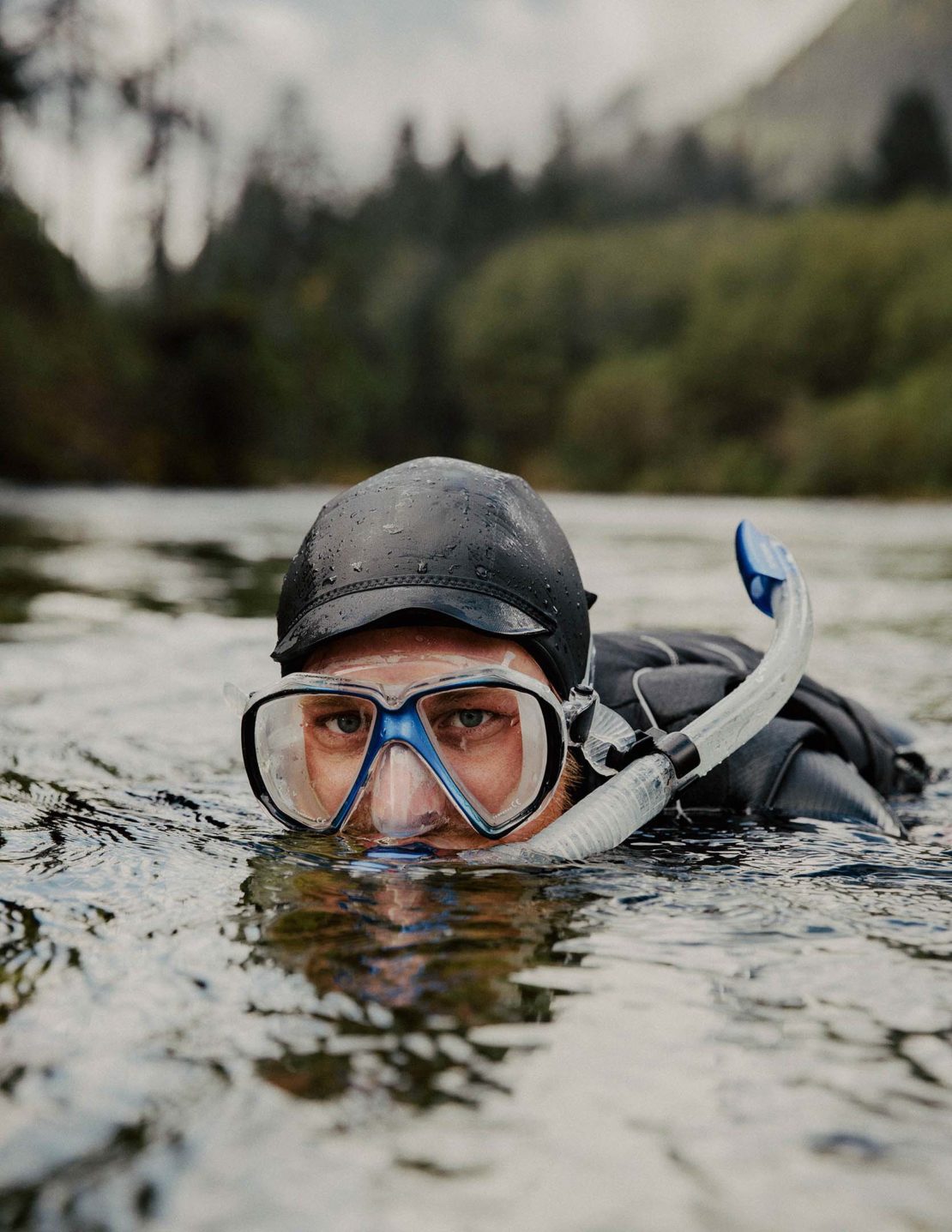 Man with snorkel and goggles in river with trees and overcast sky. The person is waist-deep in the water with only their head visible.
