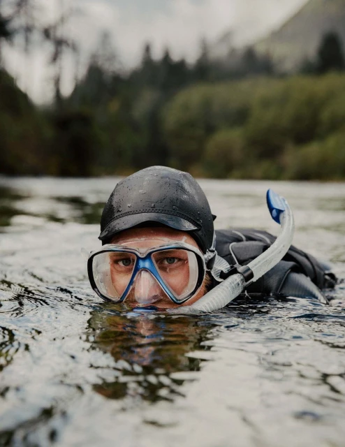 Man with snorkel and goggles in river with trees and overcast sky. The person is waist-deep in the water with only their head visible.