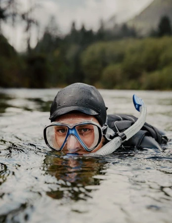 Man with snorkel and goggles in river with trees and overcast sky. The person is waist-deep in the water with only their head visible.