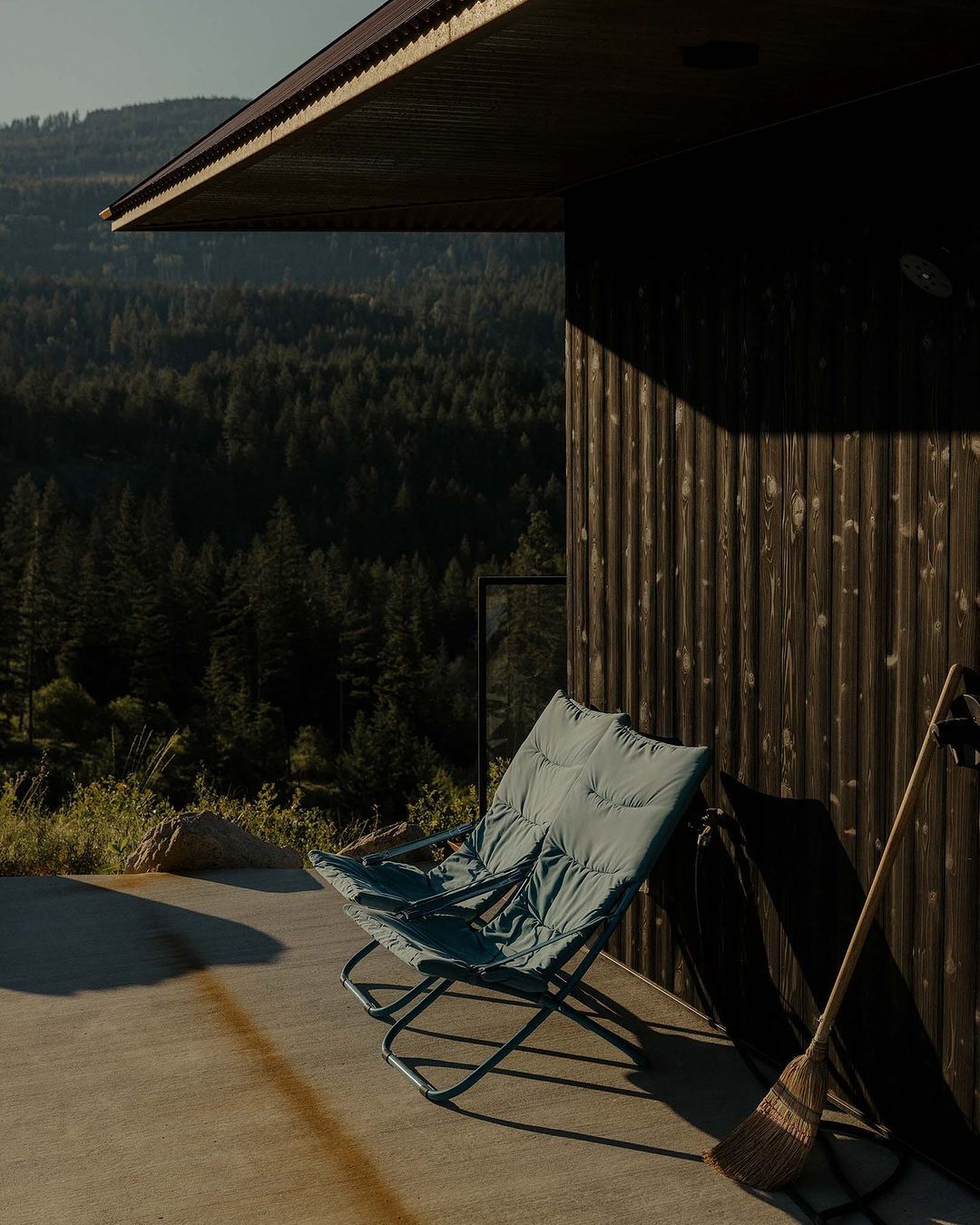 Exterior view of modern cabin with wood facade and a blue chair on the patio, with a forest in the background on a sunny day.