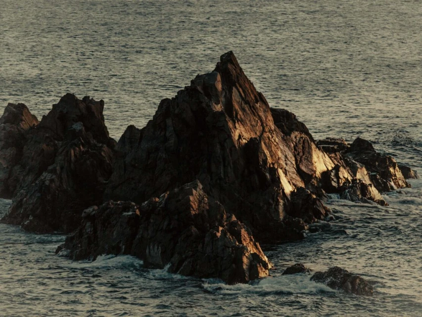 Rocky outcropping in the ocean, illuminated by the sun. Waves are gently lapping at the rocks in this serene coastal scene.