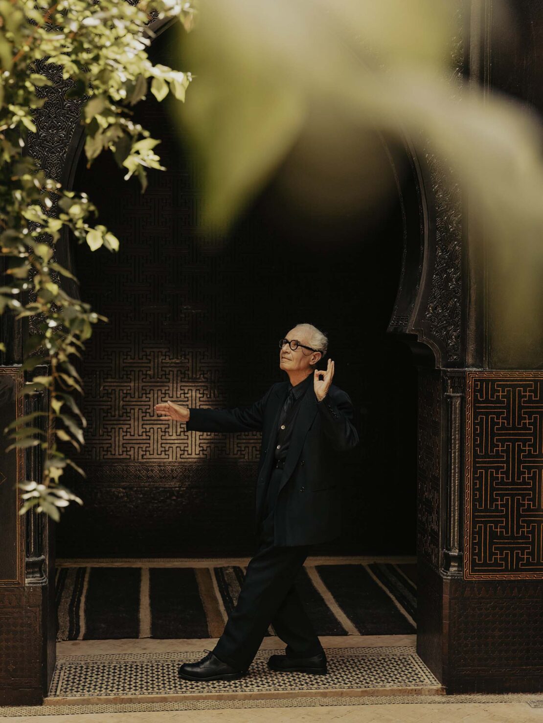 Stylish elderly man in a dark suit walks through an ornately tiled archway with lush green foliage in an outdoor setting.
