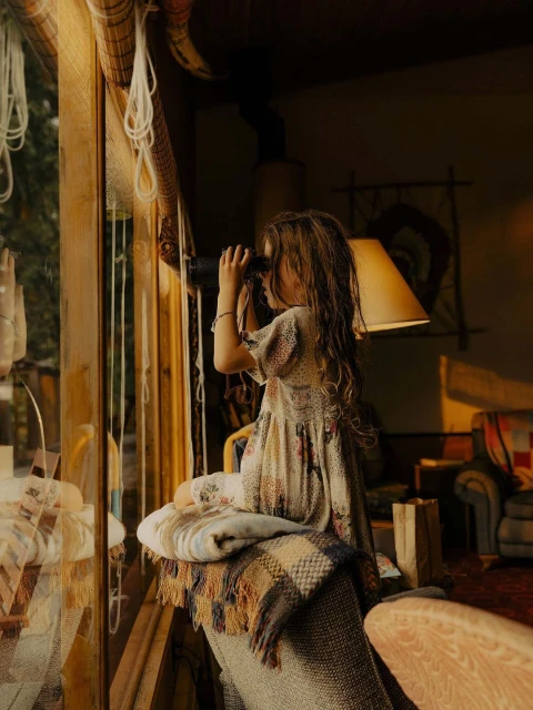 Child looks through binoculars near a window in a cozy, warm-toned interior, beside a blanket-draped chair, creating a nostalgic scene.