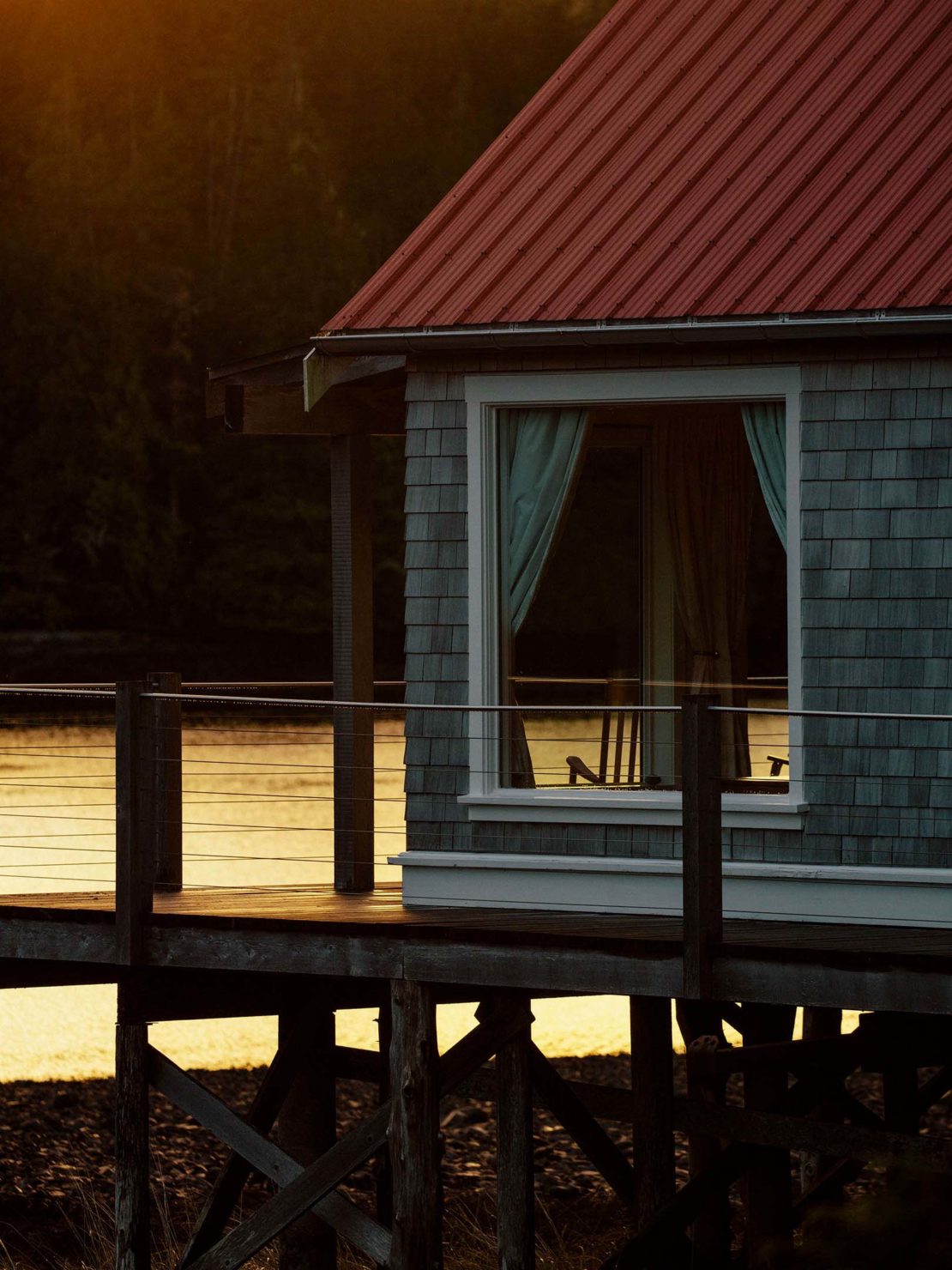 A wooden stilt house with a red roof is reflected in calm water surrounded by dark forest, creating a serene and secluded retreat.
