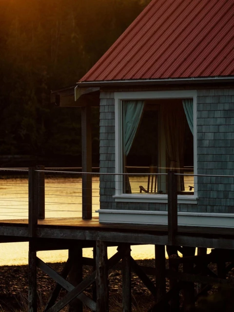 A wooden stilt house with a red roof is reflected in calm water surrounded by dark forest, creating a serene and secluded retreat.