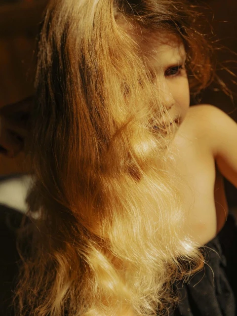 Portrait of a young girl with long blonde hair partially obscuring her face, with warm lighting and a dark background.