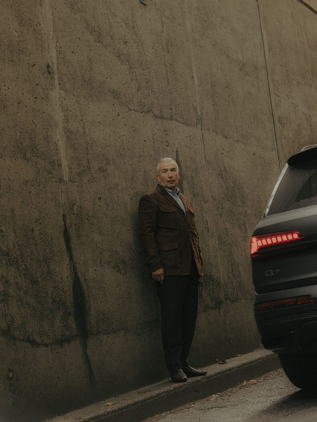 An adult male stands next to a modern SUV on an overcast day, leaning slightly against a textured concrete wall in an outdoor setting.