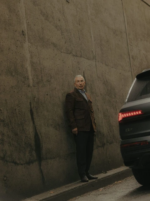 An adult male stands next to a modern SUV on an overcast day, leaning slightly against a textured concrete wall in an outdoor setting.