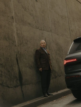 An adult male stands next to a modern SUV on an overcast day, leaning slightly against a textured concrete wall in an outdoor setting.
