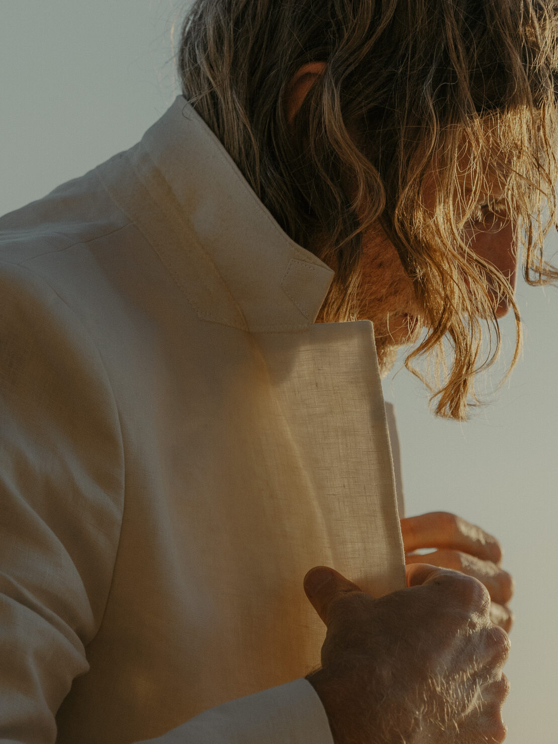 Close-up shot of a blonde adult male model wearing a light-colored linen blazer, partially open, in natural light with a warm tone.